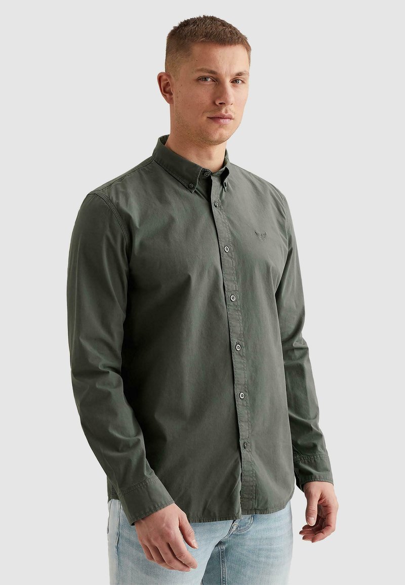 Jeune homme aux cheveux courts portant une chemise boutonnée vert foncé et un jean bleu clair, debout devant un fond clair uni.