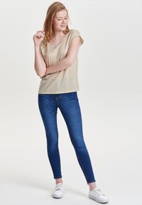 Guld, kortærmet top med V-hals; parret med blå, tætsiddende jeans og hvide sneakers. Skjorten har en subtil glans og blød tekstur.