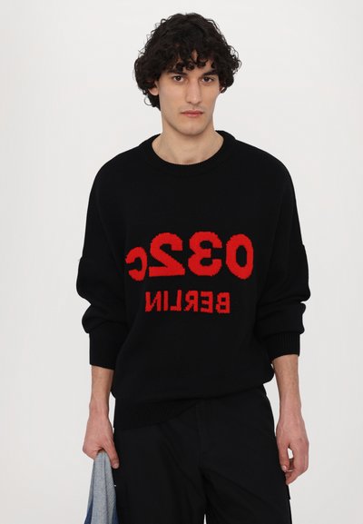 Czarny sweter dzianinowy z dużym czerwonym napisem "032 BERLIN". Żebrowany dekolt i mankiety. Model trzyma szary materiał. Casualowy design.