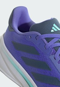 Zapatilla deportiva morada con una parte superior de malla texturizada, acentos en color azul verdoso y tres rayas oscuras a los lados. Suela blanca con un patrón de huella visible.