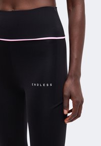 Personne à la peau foncée portant des leggings noirs taille haute avec une fine bande rose et le mot "ENDLESS" sur la cuisse.
