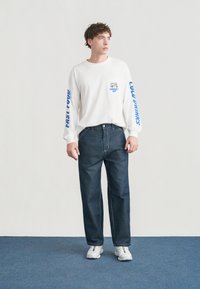 Λευκό πουκάμισο μακρυμάνικο με μπλε τύπωμα στα μανίκια και μπλε λογότυπο στην τσέπη· σκούρο τζιν παντελόνι· λευκά αθλητικά παπούτσια σε μπλε δάπεδο.