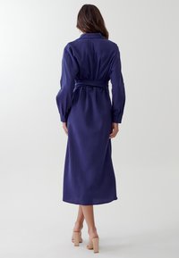Abito a maniche lunghe blu navy con vita a fascia, realizzato in tessuto liscio. Presenta un collo e polsini con bottoni, che scende fino a metà polpaccio.