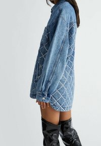 Denim shirt dress with a quilted pattern accented by silver studs. Features a button collar and long sleeves. Blue color and oversized fit.