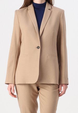 Kvinde iført en beige skræddersyet blazer og matchende bukser over en mørkeblå rullekrave, stående med hænderne afslappet langs siderne.