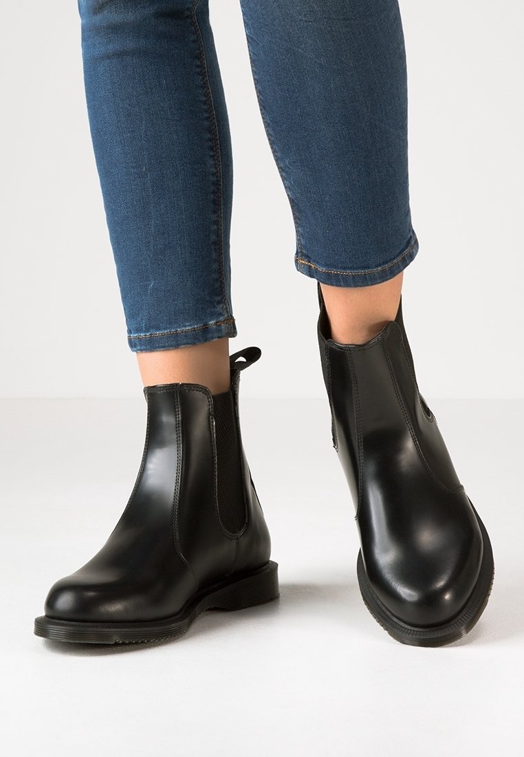 Czarne skórzane botki sięgające kostki z elastycznymi bocznymi panelami i gładkim wykończeniem, zestawione z przykrótkimi niebieskimi dżinsami. Płaskie gumowe podeszwy.