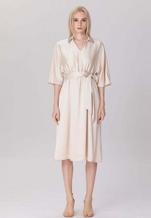 Femme blonde debout portant une robe midi en satin crème avec des manches aux coudes, une taille ceinturée et un col orné, talons neutres.