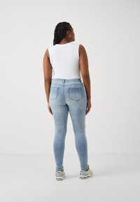 Ανοιχτό μπλε τζιν skinny παντελόνι με πίσω τσέπες, συνδυασμένο με λευκή αμάνικη μπλούζα και γκρίζα αθλητικά παπούτσια, απέναντι από έναν απλό φόντο.