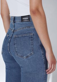 Džíny z modrého denimu s vysokým pasem a přiléhavým střihem, s výraznými zadními kapsami a černou etiketou, na které je nápis "DRDENIM."