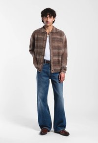 Cardigan rayé marron en matière douce, porté sur un débardeur blanc. Associé à un jean pattes d53uf bleu clair et des chaussures marron.