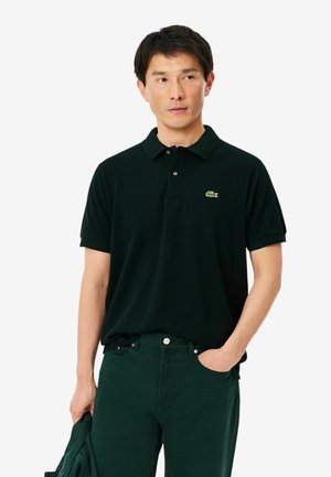 Uomo che indossa una polo nera con un logo coccodrillo verde, pantaloni verde scuro, tiene una giacca verde scuro in una mano.
