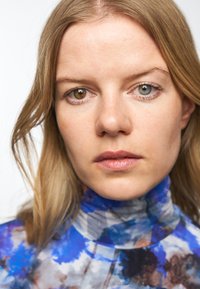 Nærbilde av en person med middels langt blondt hår, iført en høyhalset, flerfarget topp med blå og grå mønster.