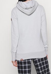 Ljusgrå huvtröja med dragsko, ribbade ärmslut och nederkant. Har ett brunt logopatch på den nedre sidan. Bärs med rutiga byxor.