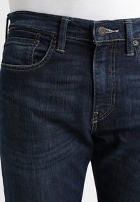 Vaqueros de mezclilla azul oscuro con un diseño clásico, que presentan un cierre de botón, cinco bolsillos y costuras contrastantes en amarillo.