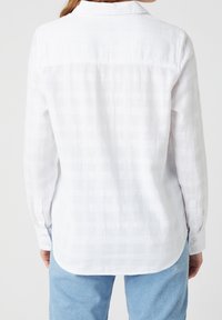 Vit långärmad skjorta med ett strukturerat rutmönster och spetsig krage. Har knäppte ärmslut och en rundad nederkant bak.