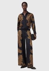 Mustast satiinist komplekt, millel on kuldne mustriline disain, koosneb pikkade varrukatega särgist ja laiades sääreosades pükstest, täiustatud musta kotiga.