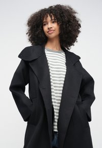 Sort uldfrakke med stor krave, løs pasform og sidelommer, lagdelt over en gråstribet skjorte, kombineret med blå jeans.