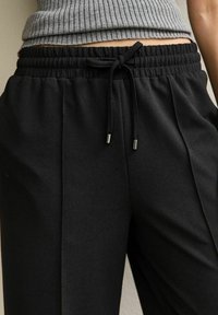 Zwarte broek met een gestructureerde afwerking, voorzien van een elastische tailleband en een trekkoord met metalen eindstukjes. Het ontwerp bevat voorplooien.
