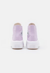 Lilac canvas high-top sneakers with white rubber soles, featuring circular black and white logo patches on the outer ankle and textured detailing.