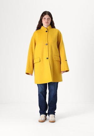 Geel wollen jas met een hoge kraag, twee voorkruiszakken en drie knopen. Losse pasvorm, gecombineerd met blauwe spijkerbroeken en beige sneakers.
