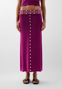 Gonna lunga e aderente di colore viola con una cintura decorativa di perline. Presenta una fila di bottoni a forma di conchiglia lungo la parte anteriore. Indossata con sandali marroni con fibbia.