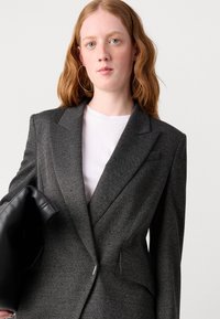 Blazer en chevrons gris avec une coupe ajustée, des revers structurés et deux poches avant, associé à un t-shirt blanc. Objet en cuir noir tenu.