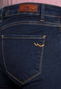 Jeans de mezclilla azul oscuro con un parche de cuero marrón etiquetado "LTB". Presenta un diseño de costura distintivo y un pequeño diseño de ola bordado en el bolsillo.