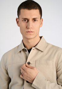 Giacca di cotone beige con chiusura frontale a bottoni e tasca sul petto. Colletto corto e dettagli di cucitura visibili sul tessuto.