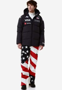 Giacca a vento nera con cappuccio, con loghi. Pantaloni colorati con motivo a stelle e strisce rosse, bianche e blu. Calzature nere.