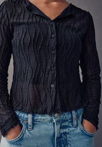 Schwarze, durchsichtige Bluse mit strukturiertem, wellenförmigem Muster und Knopfverschluss. Lange Ärmel mit gerafften Details. Kombiniert mit hellblauen Denim-Jeans.