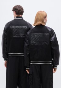Jachete bomber negre cu mâneci din piele și manșete rib, având un panou texturat negru cu inscripția "TOMMY JEANS" în relief pe spate.