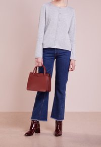 Frau in grauem, geknöpftem Pullover, blauen Jeans und dunkelroten Stiefeletten, die eine passende dunkelrote Handtasche vor einem hellen Hintergrund hält.