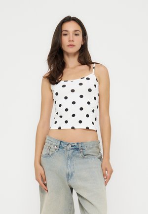 Jeune femme aux longs cheveux foncés portant un débardeur court blanc à pois noirs et un jean taille haute bleu clair sur un fond uni.
