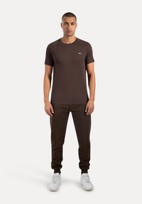 Camiseta marrón de manga corta con cuello redondeado, combinada con pantalones jogger marrones a juego y zapatillas blancas. Diseño simple y ajustado.