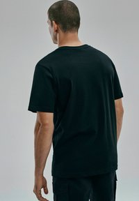 Czarna bawełniana koszulka z krótkim rękawem, okrągłym dekoltem, luźnym krojem i gładką fakturą, pokazana z tyłu na neutralnym tle.