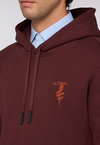 Maroon mikina s kapucí a stahovacími šňůrkami, s malým oranžovým vyšitým designem. Má texturovanou tkaninu a vrstvený límec košile pod ní.