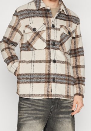 Man draagt een beige, bruin en grijs geruit wollen overhemd met borstzakken en vervaagde zwarte jeans, staand tegen een effen achtergrond.