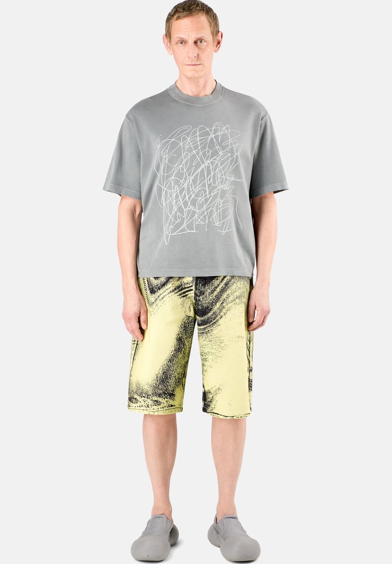 Camiseta de algodón gris con diseño gráfico abstracto blanco, combinada con pantalones cortos amarillos hasta la rodilla que presentan patrones ondulados negros y costuras visibles.