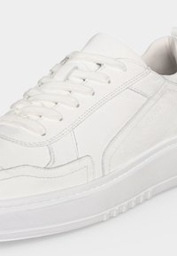 Baskets en cuir blanc avec semelle épaisse, lacets à l'avant et détails en panneaux texturés, présentées en angle de côté sur un fond clair.