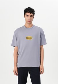 Grå t-skjorte med kort erme laget av bomull, med en gul oval "GUESS" logo på brystet. Enkel, avslappet passform.
