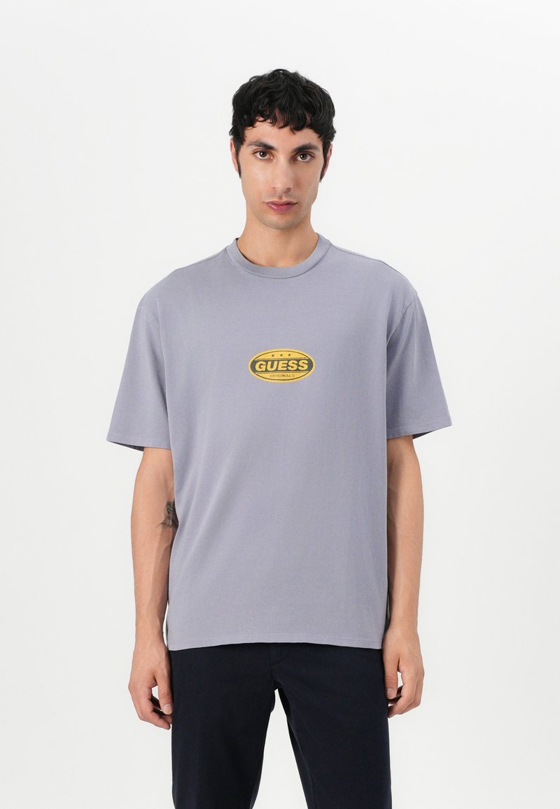 Grå t-skjorte med kort erme laget av bomull, med en gul oval "GUESS" logo på brystet. Enkel, avslappet passform.