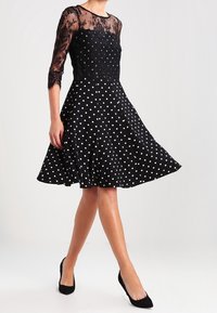 Svart klänning med prickar, en åtsittande bodice av spets och trekvartslånga ärmar. Urtstlängd kjol, slät textur, och bärs med svarta skor.