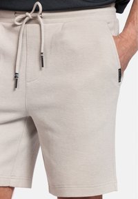 Lichtbeige shorts van textuurstof, met een trekkoord in de taille en metalen uiteinden, en zijzakken. Subtiele merknaam aan de zijkant.
