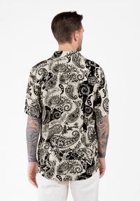 Hombre con el cabello corto y castaño y brazos tatuados, vistiendo una camisa de manga corta beige con un patrón paisley en blanco y negro, de pie de espaldas.