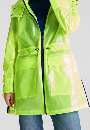 Impermeable amarillo neón semi-transparente con cremallera frontal, cintura ajustable, bolsillos con solapa y botones a presión en las mangas, llevado sobre vaqueros azules.