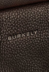 Cuir texturé marron foncé avec une finition en relief, arborant un logo "BURKELY" en relief. Le gros plan met en avant le grain du matériau.
