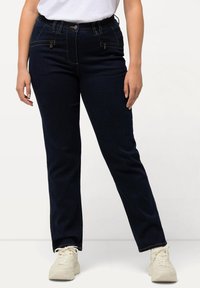 Jeans de mezclilla azul oscuro con un corte ajustado, que presentan una textura suave, una cinturilla elástica y bolsillos frontales con cremallera. Completados con zapatillas de color claro.