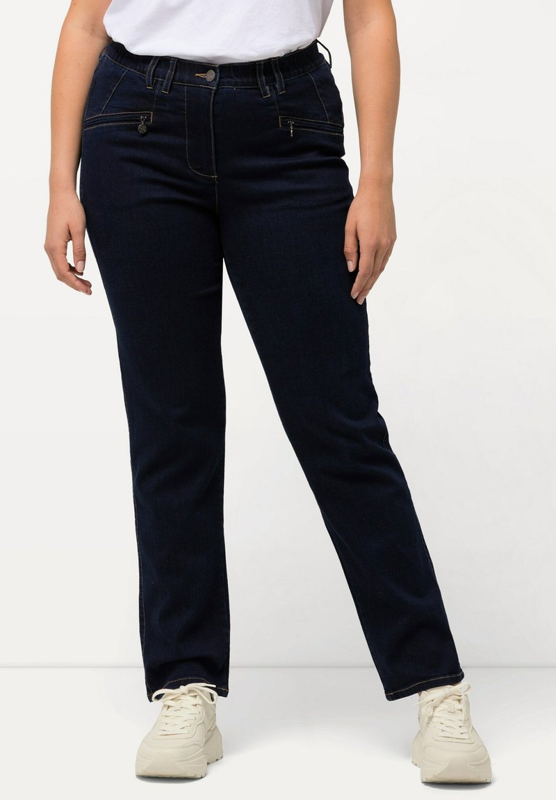 Jeans de mezclilla azul oscuro con un corte ajustado, que presentan una textura suave, una cinturilla elástica y bolsillos frontales con cremallera. Completados con zapatillas de color claro.