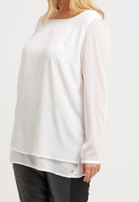 Blusa blanca de manga larga con escote redondeado, que presenta mangas transparentes y un cuerpo liso, combinada con pantalones de cuero negros.