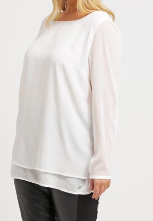 Haut blanc à manches longues avec un col rond, présentant des manches transparentes et un corps uni, associé à un pantalon en cuir noir.